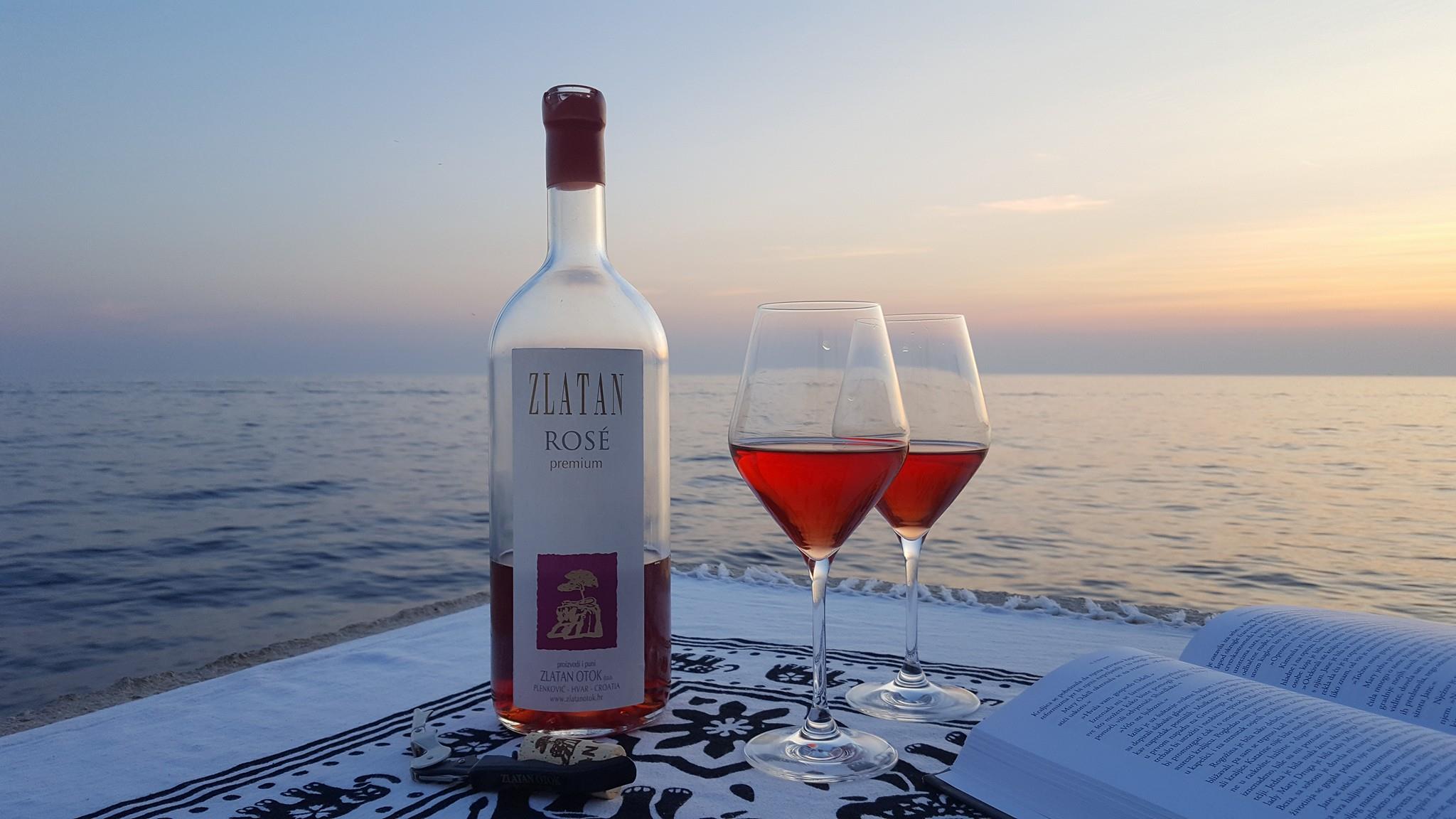 Cette image montre une bouteille à moitié vide de vin rose Zlatan Otok, deux verres pleins de rose et un livre ouvert avec la mer et le coucher de soleil inférieur. 