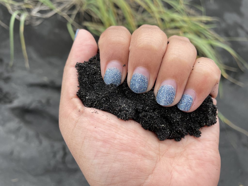 Une main aux ongles bleus brillants tient une poignée de sable noir brillant.
