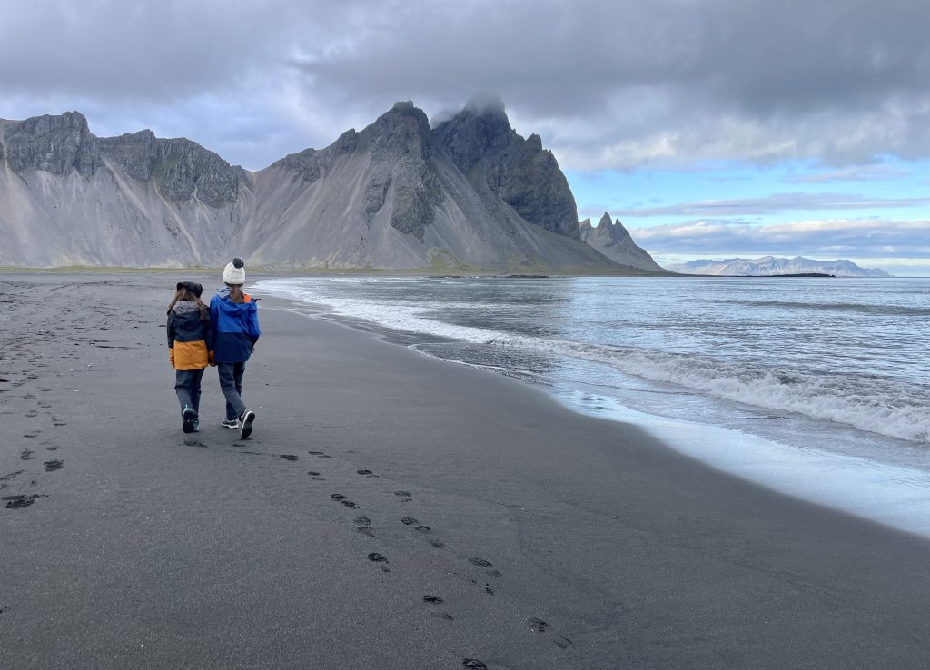 Deux enfants habillés en hiver marchent au bord de l’eau, derrière eux des montagnes déchiquetées.