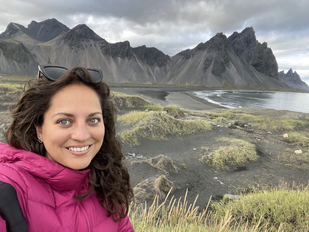 Kate portait un manteau rose avec de longs cheveux noirs ondulés et souriait pour le selfie. Derrière elle se trouvaient les collines noires et abruptes du Vestrahorn.