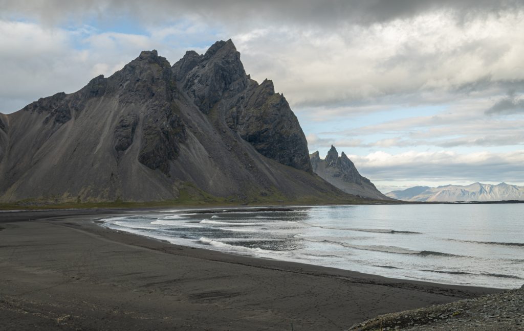 La plage noire se trouve à côté des eaux calmes, avec une montagne noire et déchiquetée en arrière-plan.